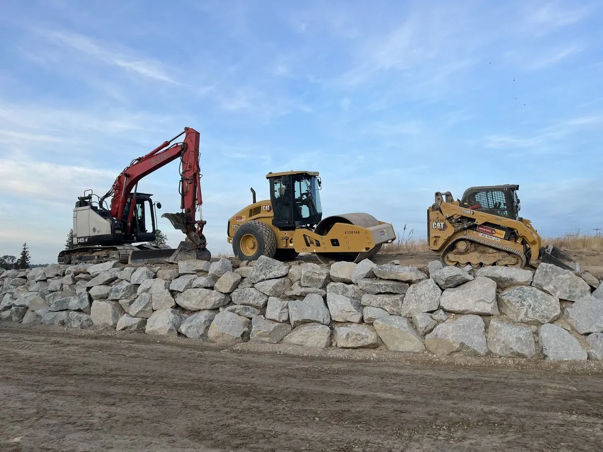 Excavator, smooth drum roller and compact track loader lined up behind a newly built boulder retaining wall