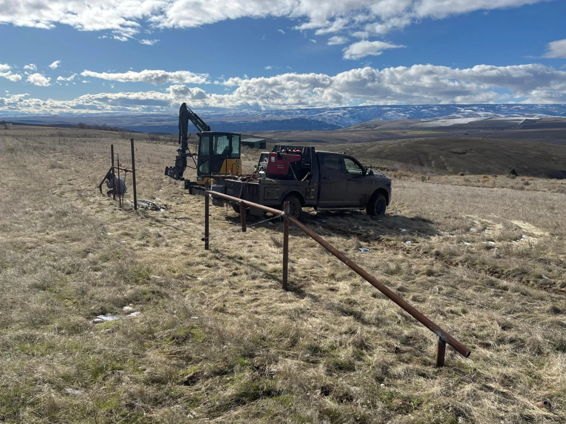 Steel pipe fence posts and rails being installed on open rangeland, with a compact excavator and a welding truck behind
