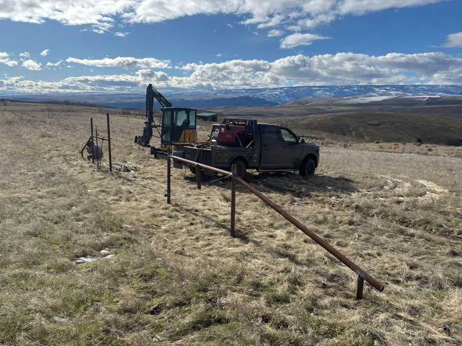 Steel pipe fence posts and rails being installed on open rangeland, with a compact excavator and a welding truck behind